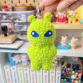 Green alien plush keychain held in front of a shelf with various items