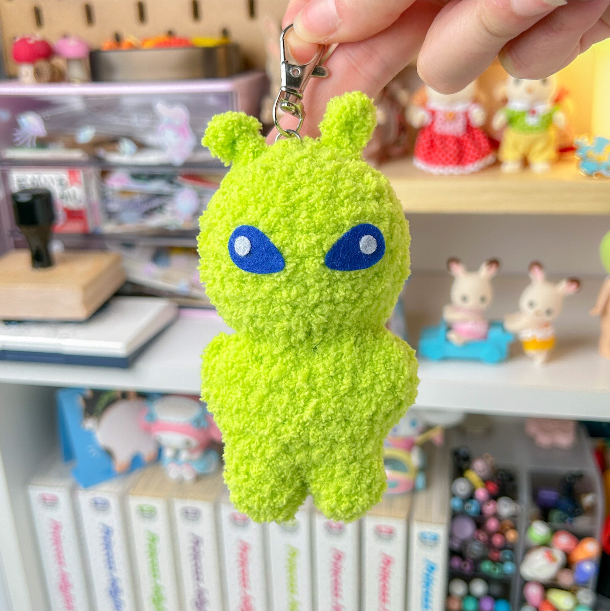 Green alien plush keychain held in front of a shelf with various items