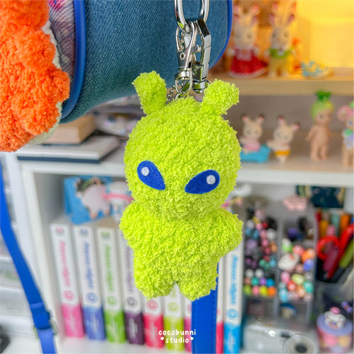 Green alien keychain hanging from a blue bag with a shelf in the background