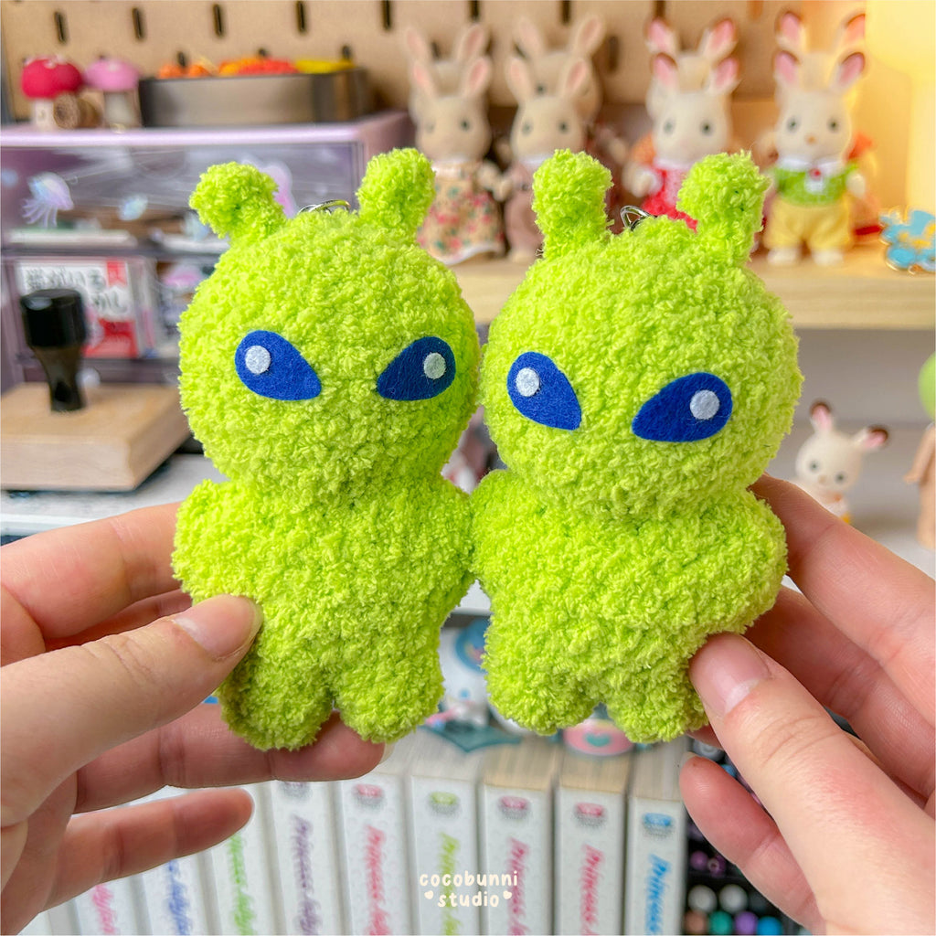 Two green plush toys with blue eyes held by a person, with a blurred background of shelves and toys.