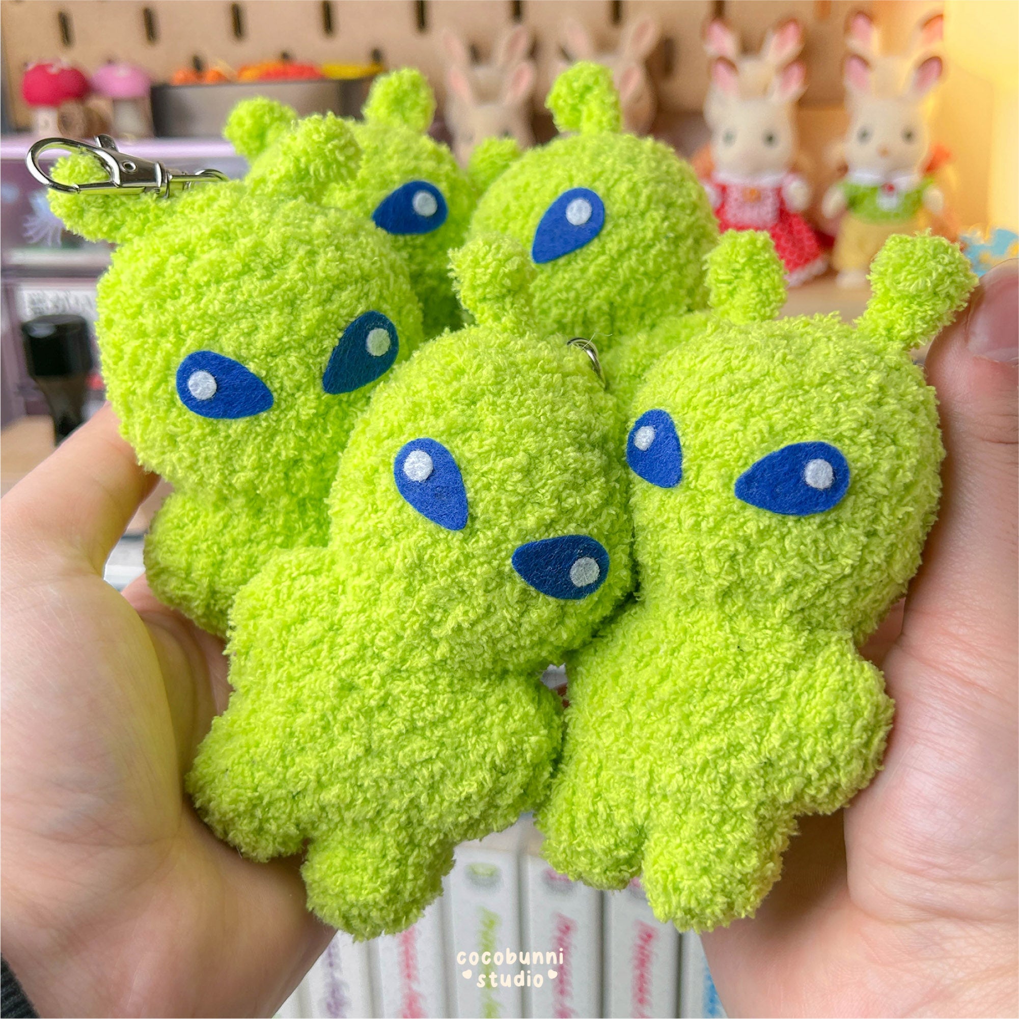 Green plush keychains with blue eyes held by a hand, with a blurred background.