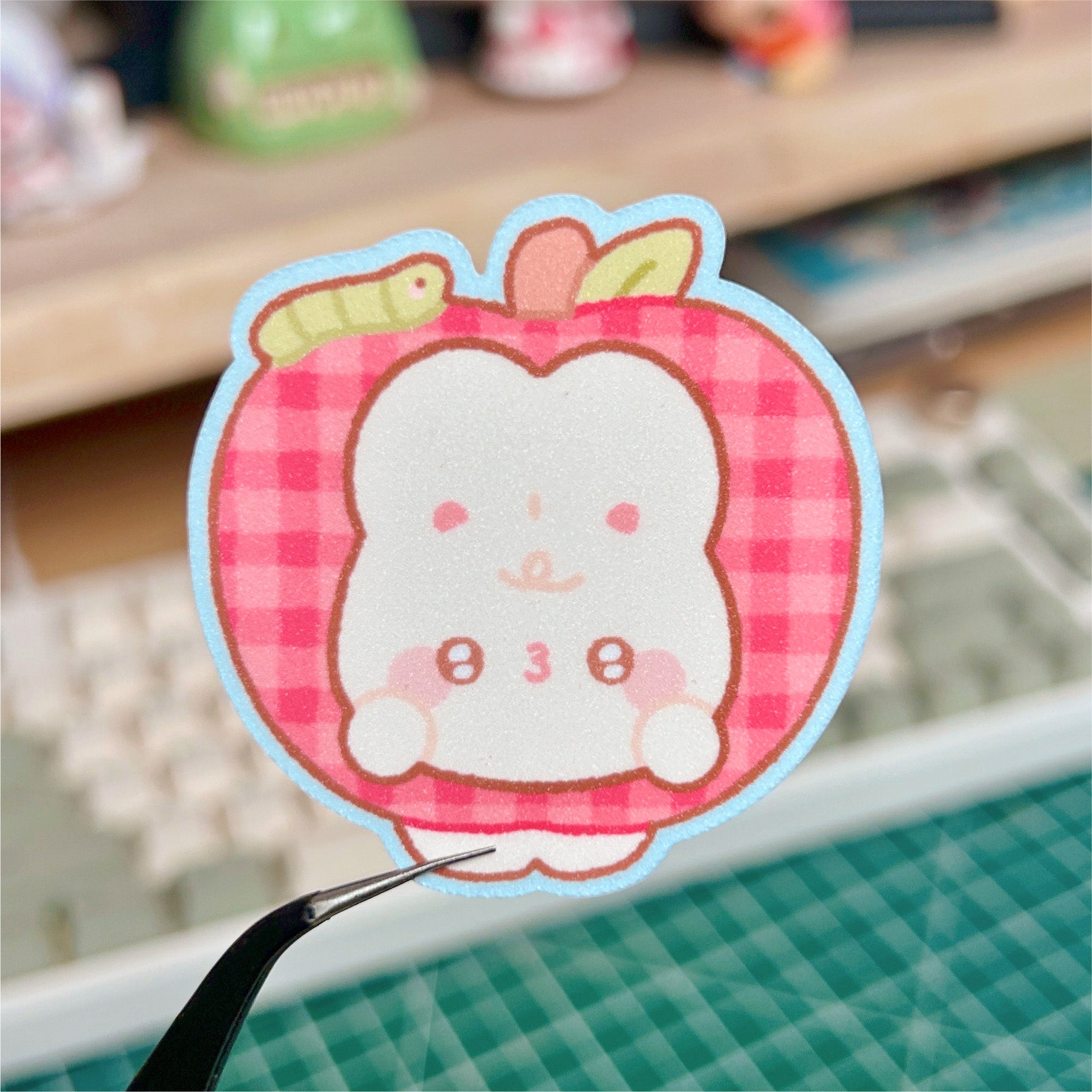 Sticker of a cartoon character in a pink checkered frame held by tweezers.