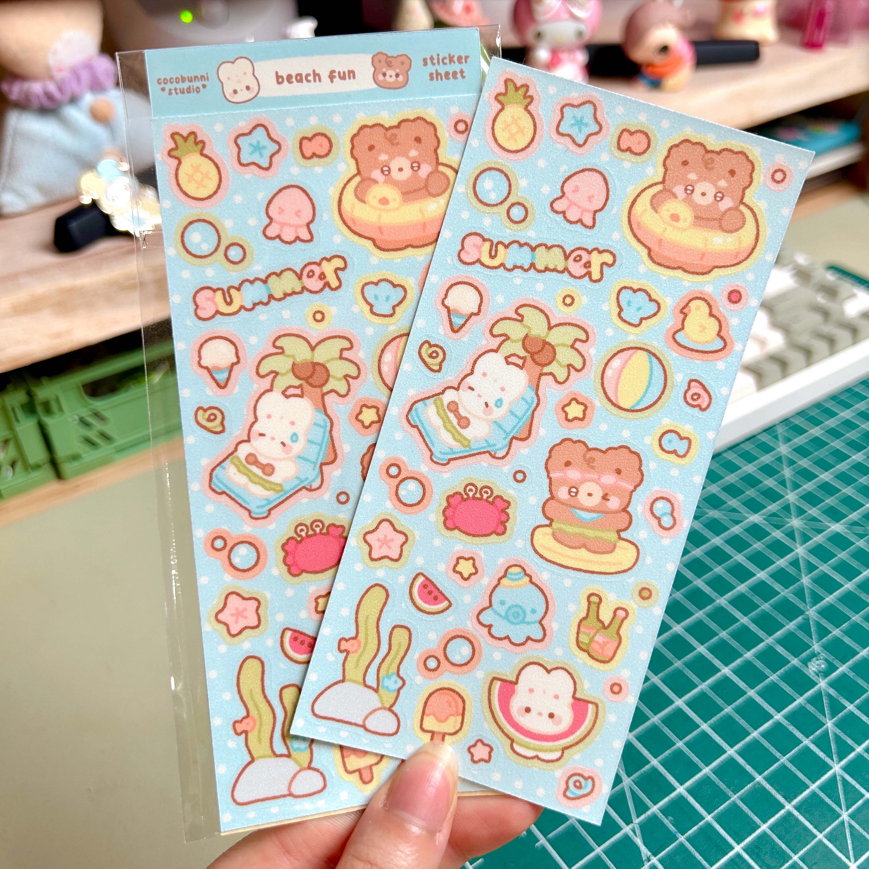 Two sheets of colorful stickers with beach-themed designs held by a hand on a cutting mat.