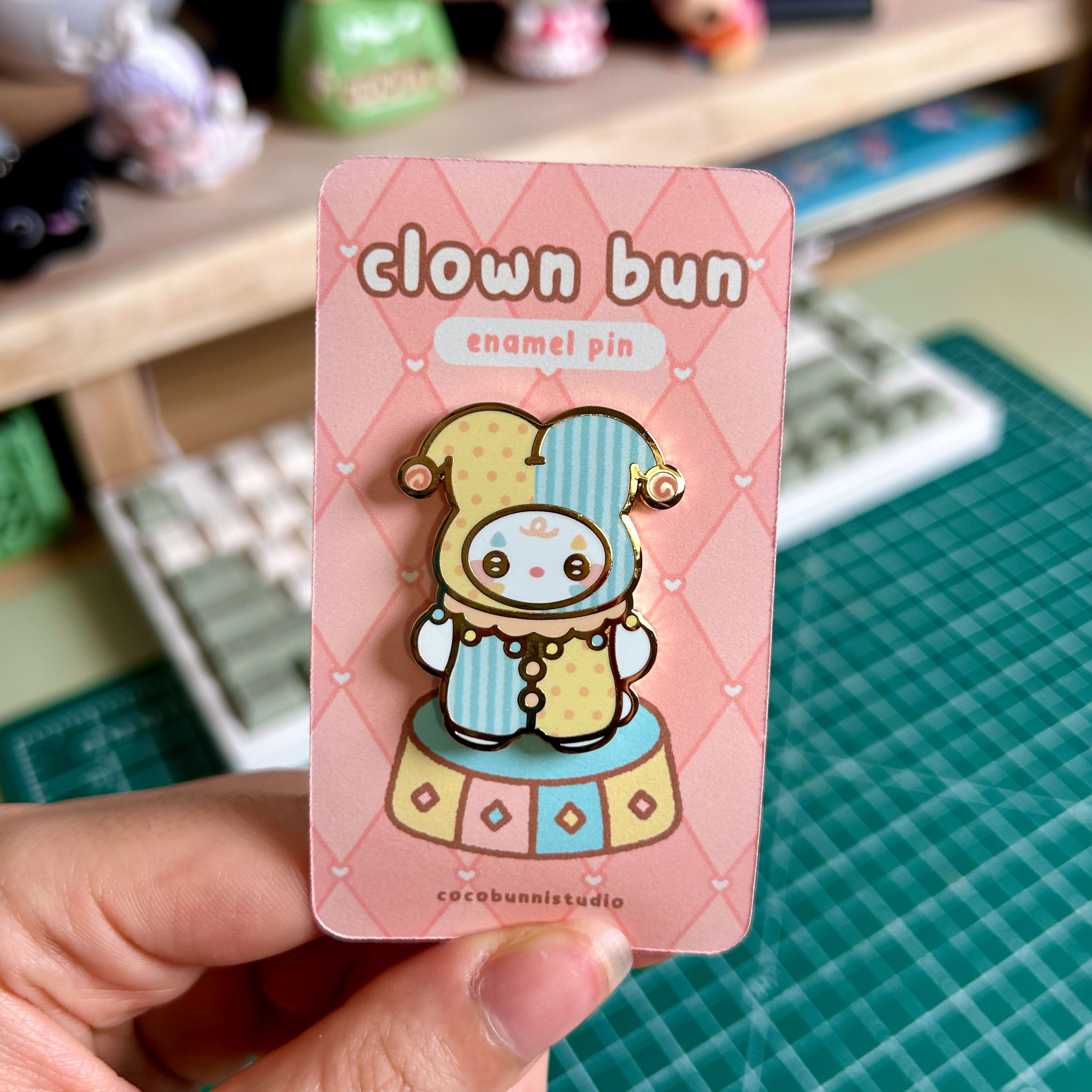 Clown bun enamel pin on a pink card held by a hand with a desk and keyboard in the background.