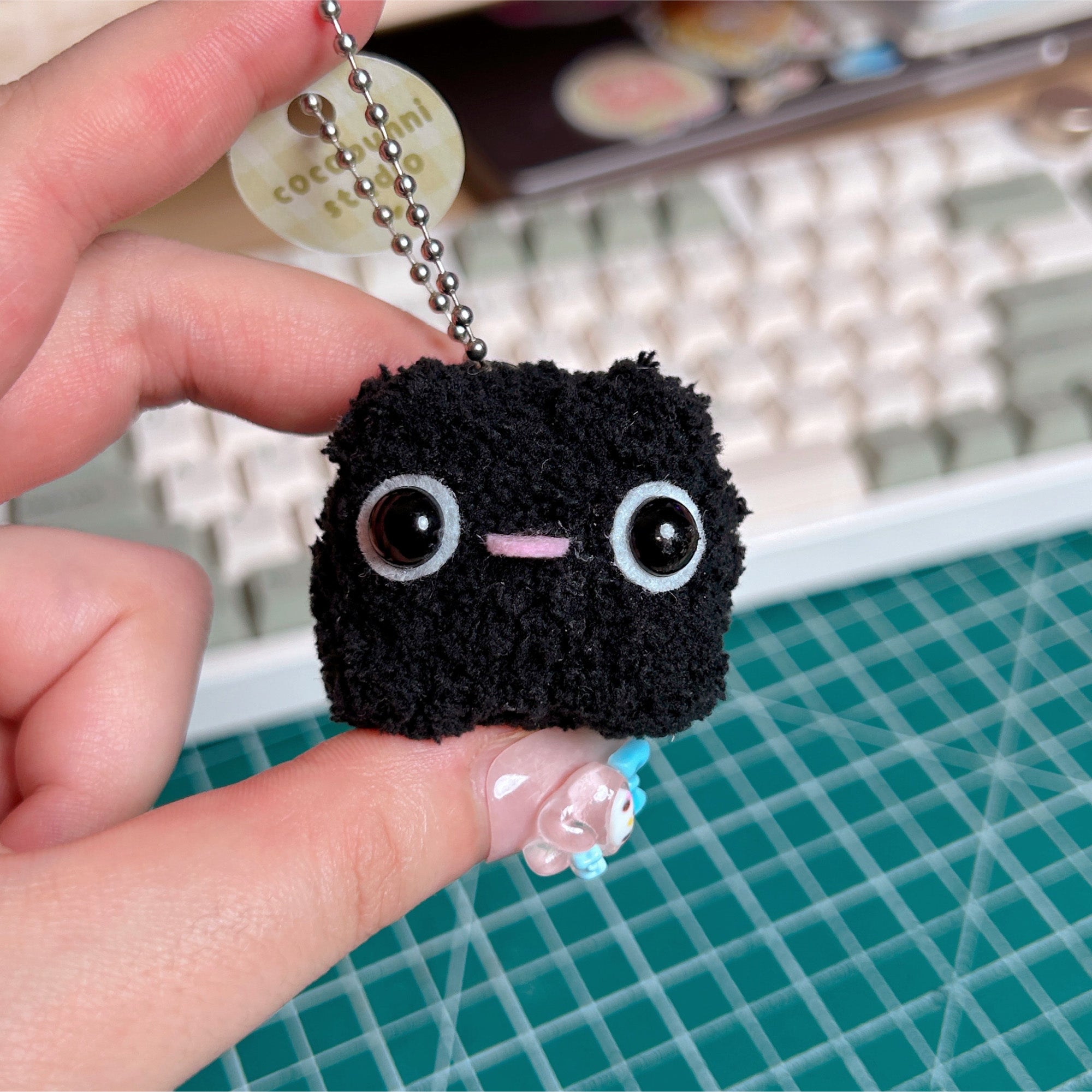 Black plush keychain held by a hand on a grid surface.