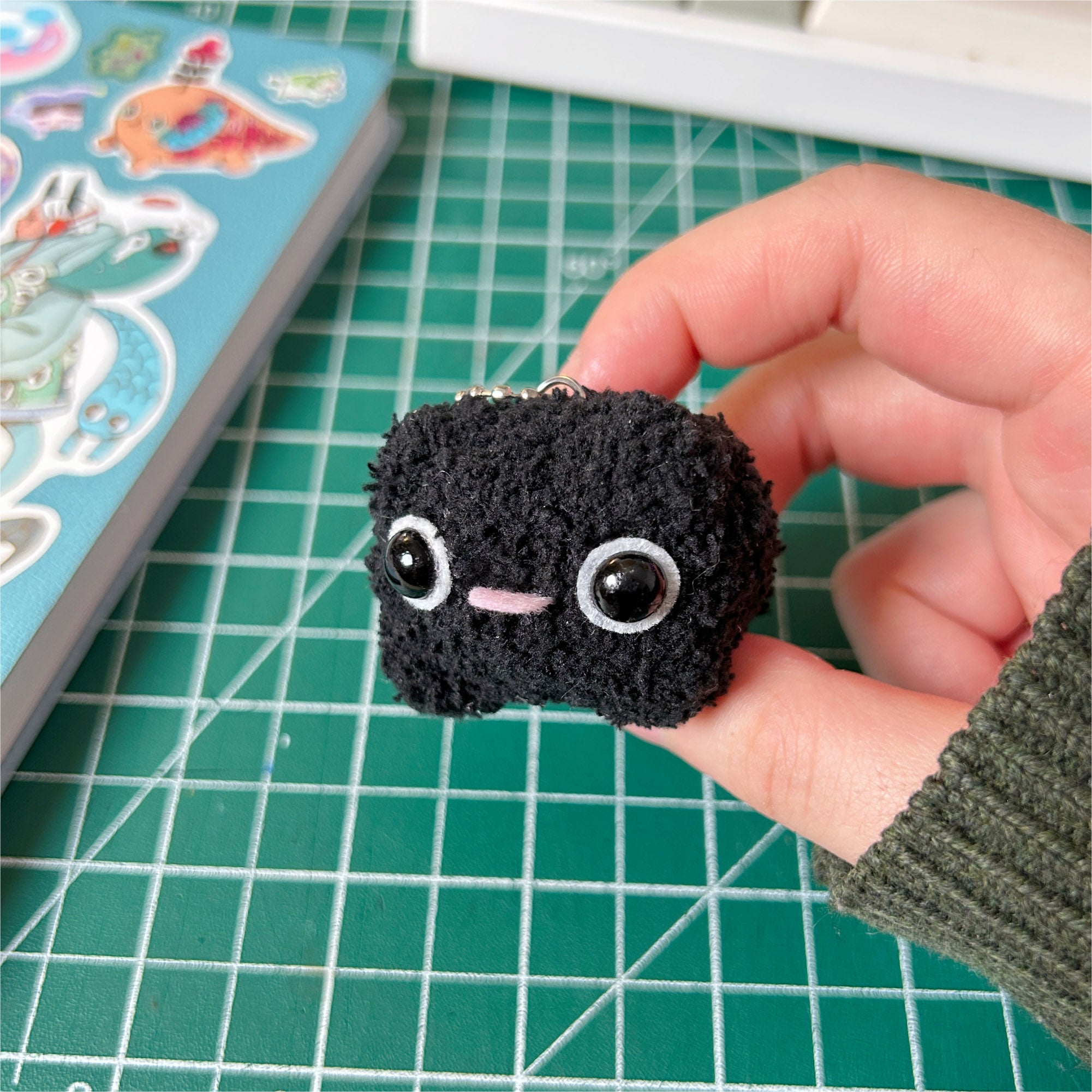 Small black plush toy held by a hand on a green cutting mat.