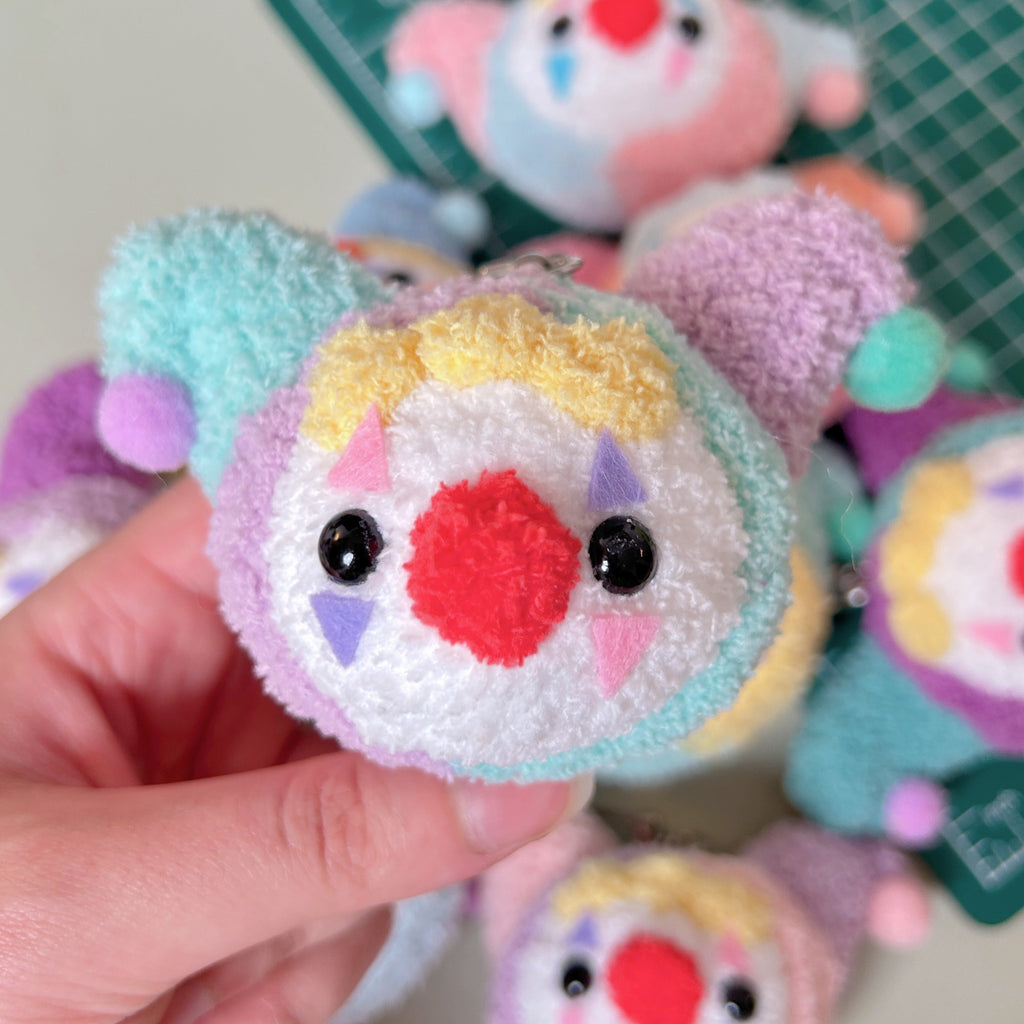 Colorful plush toy with a round face held by a hand, with more toys in the background.