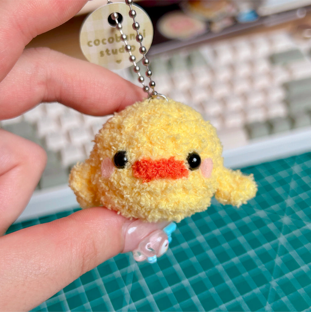 Yellow plush keychain with an orange mouth held by a hand on a green cutting mat.
