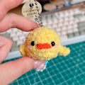 Yellow plush keychain with an orange mouth held by a hand on a green cutting mat.