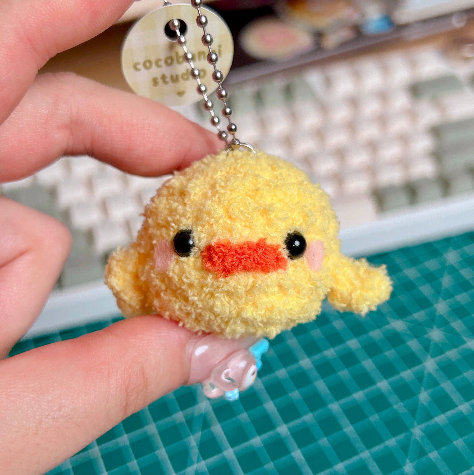 Yellow plush keychain with an orange mouth held by a hand on a green cutting mat.