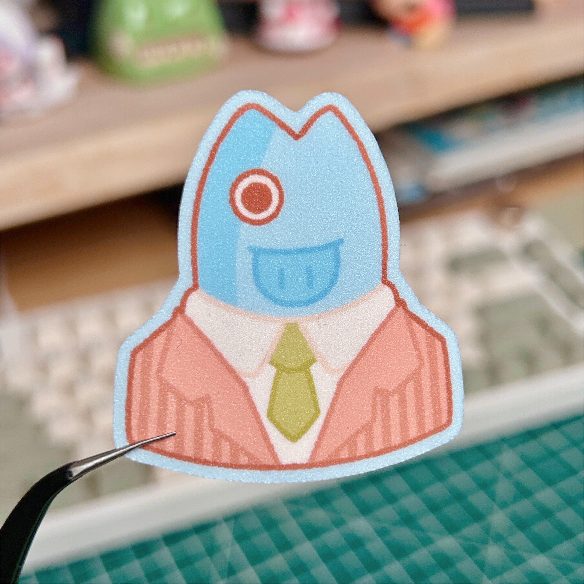 Sticker of a character in a suit with a blue head and green tie on a blurred desk background.