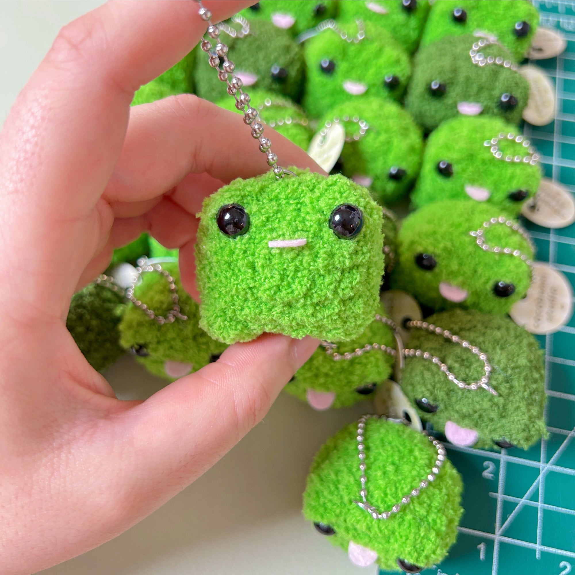 Green plush toy with a face held by a hand, with more toys in the background on a cutting mat.