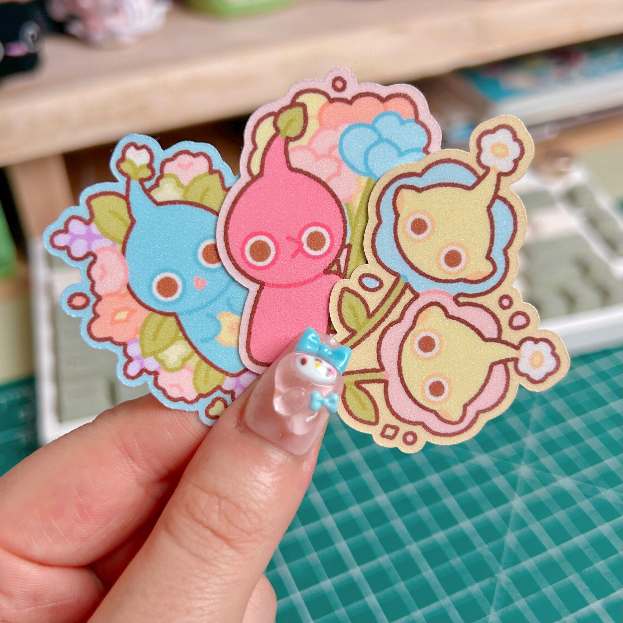 Three colorful cartoon stickers held by a hand on a green cutting mat.