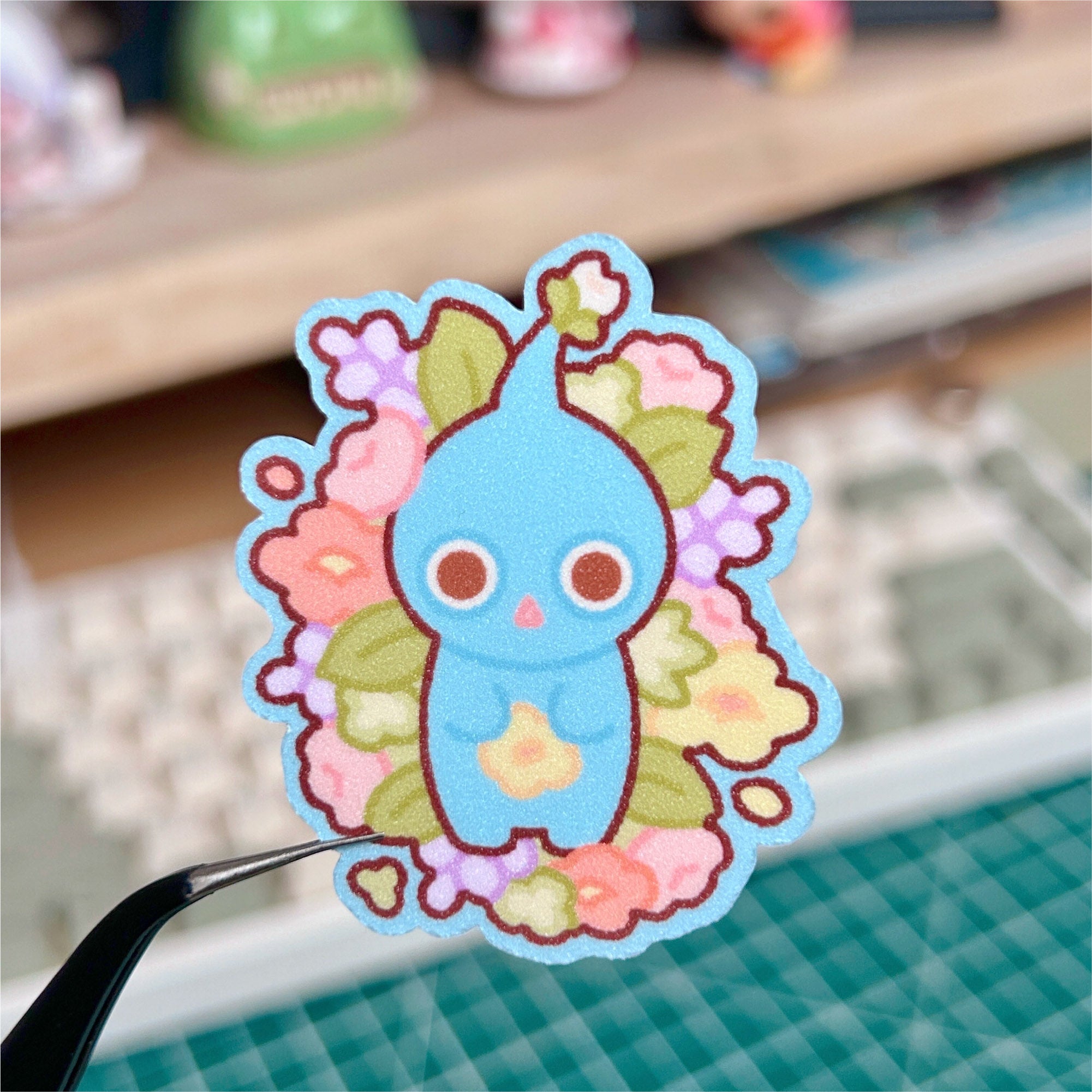 Cartoon character sticker with floral design held by tweezers on a blurred background