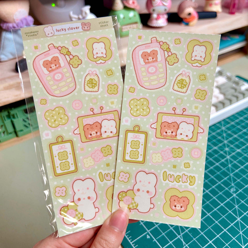 Two sheets of cute cartoon-themed stickers held by a hand on a green cutting mat.