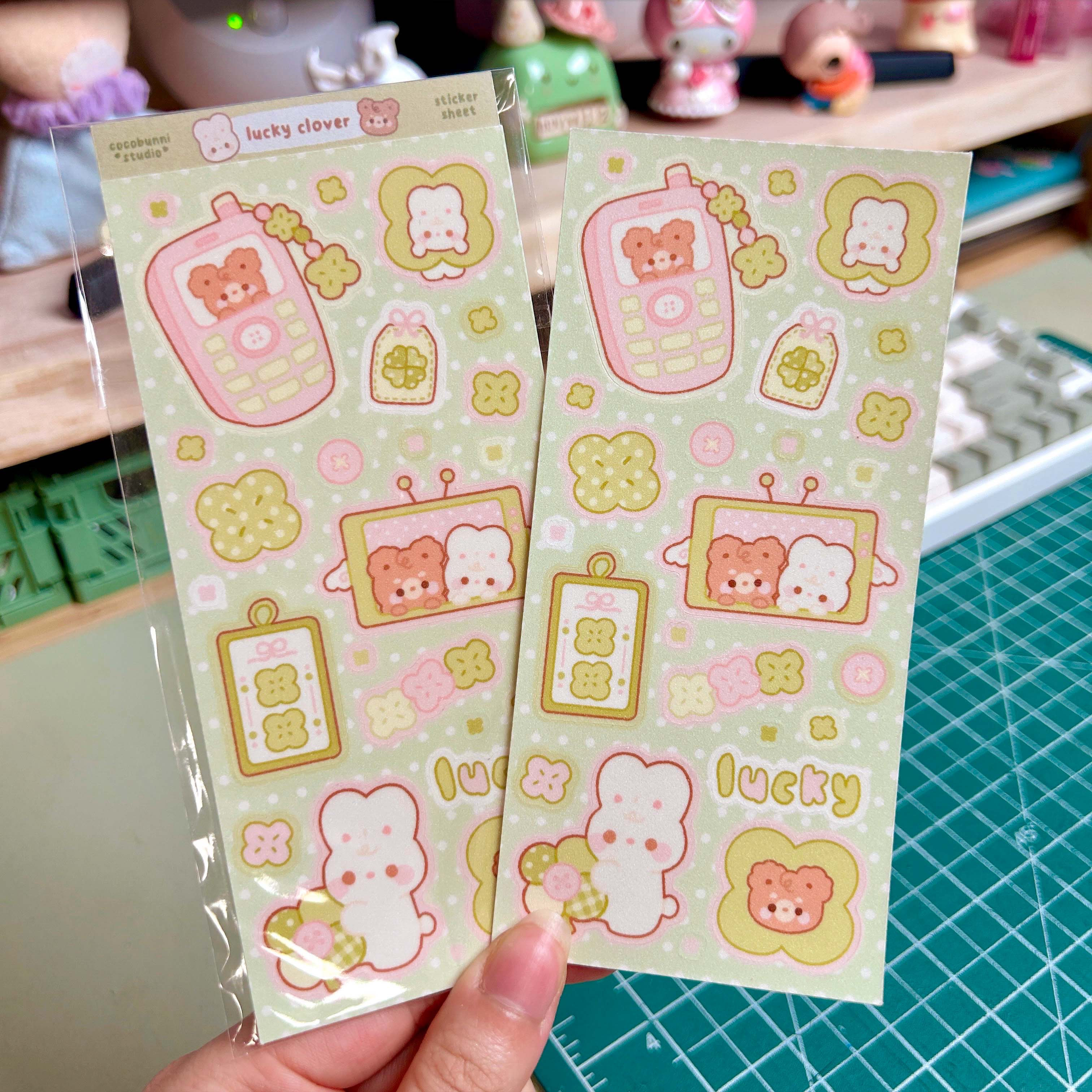 Two sheets of cute cartoon-themed stickers held by a hand on a green cutting mat.