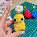 Hand holding a small yellow plush toy with a white flower on a green cutting mat.
