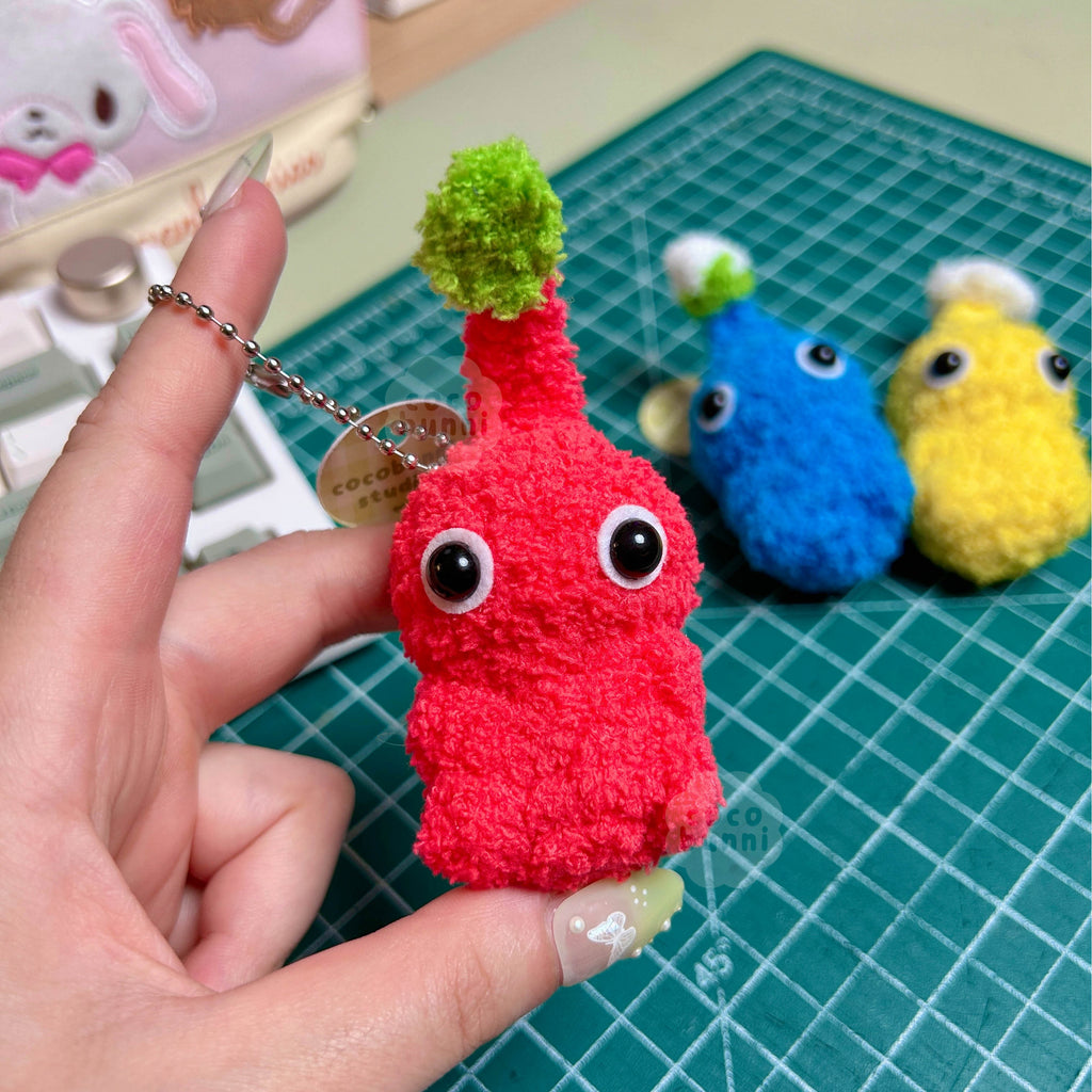 Red plush toy with a green leaf held by a hand on a cutting mat with other colorful toys.