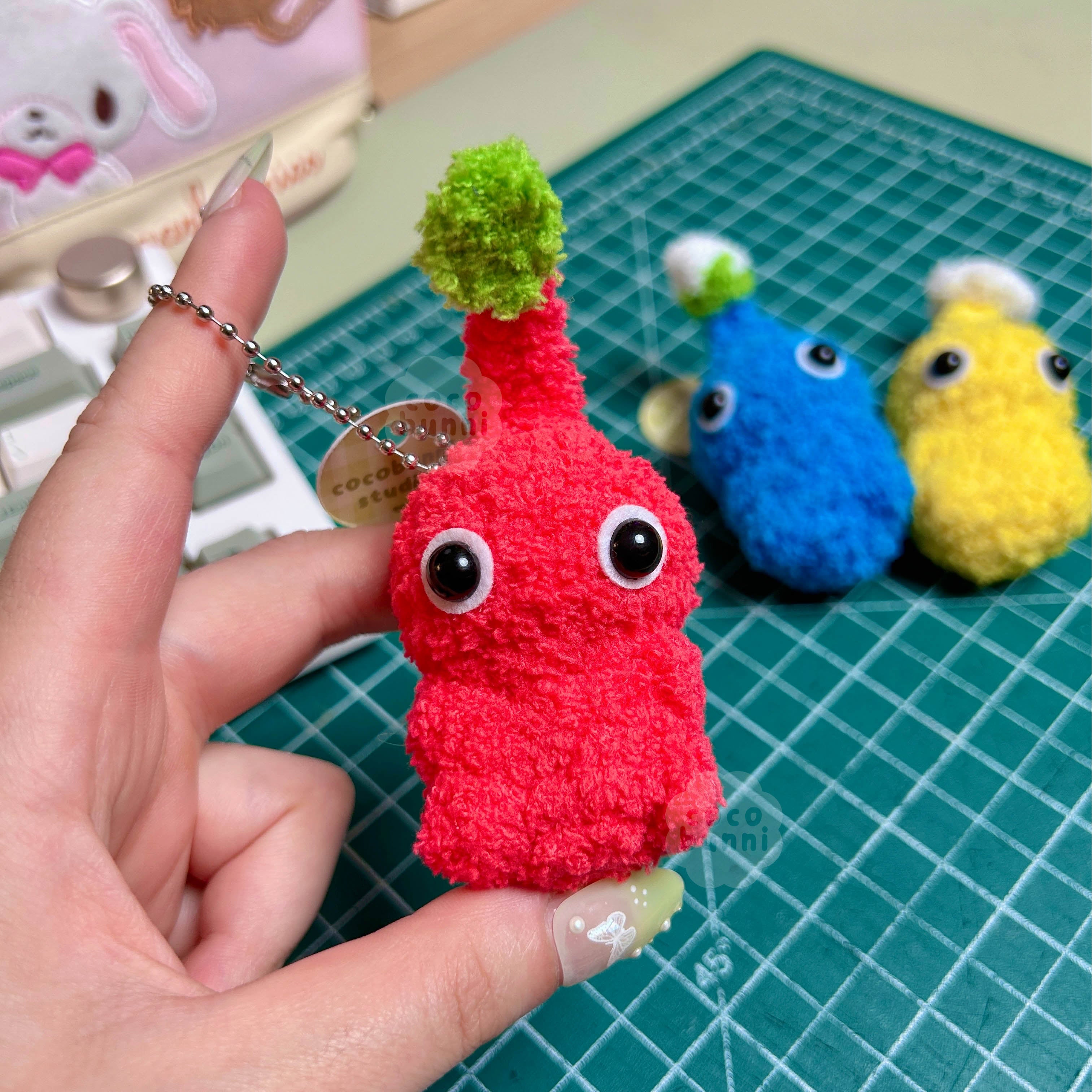 Red plush toy with a green leaf held by a hand on a cutting mat with other colorful toys.