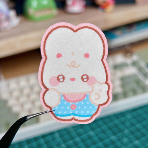 Cartoon character sticker held by tweezers on a desk with keyboard and blurred background