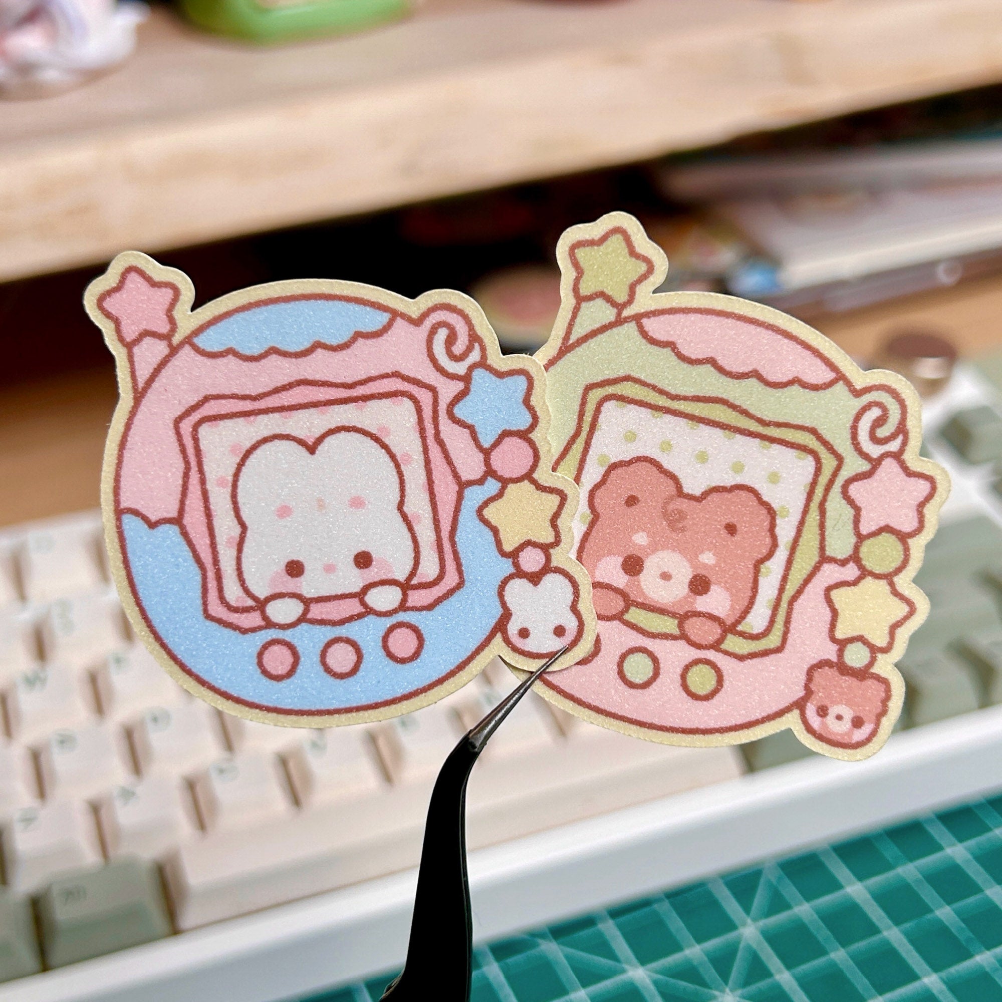 Two cartoon character stickers held by tweezers with a keyboard and cutting mat in the background.
